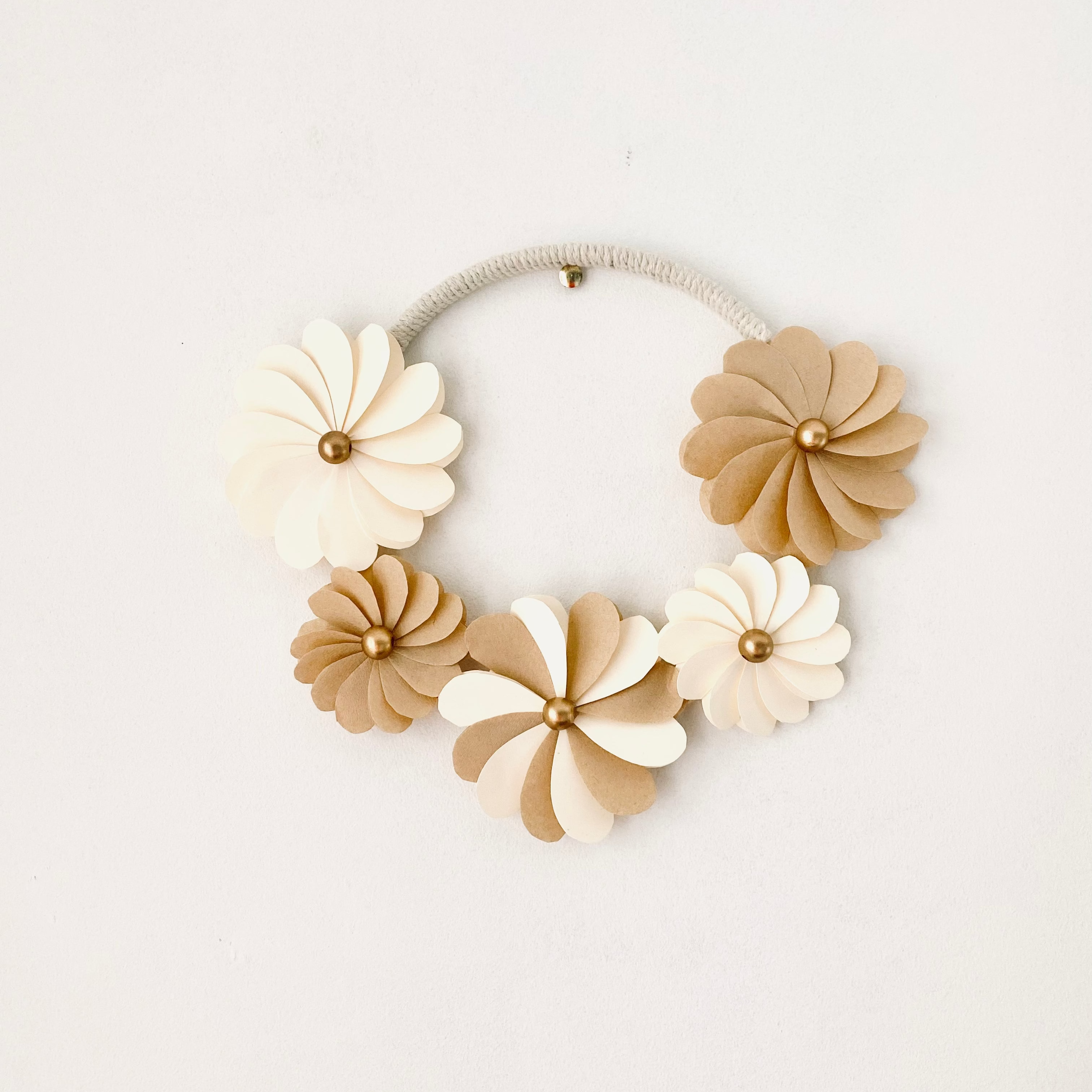 Couronne florale artisanale de 20 cm réalisée à la main. Composée de fleurs en papier écru et en filtres à café découpés un à un, chaque pétale forme une création délicate et unique. Les centres des fleurs sont ornés de perles de bois cuivrées. Cette décoration murale bohème apporte une touche douce et naturelle à un intérieur, une chambre ou un événement. Pièce unique idéale pour une décoration raffinée. ninona