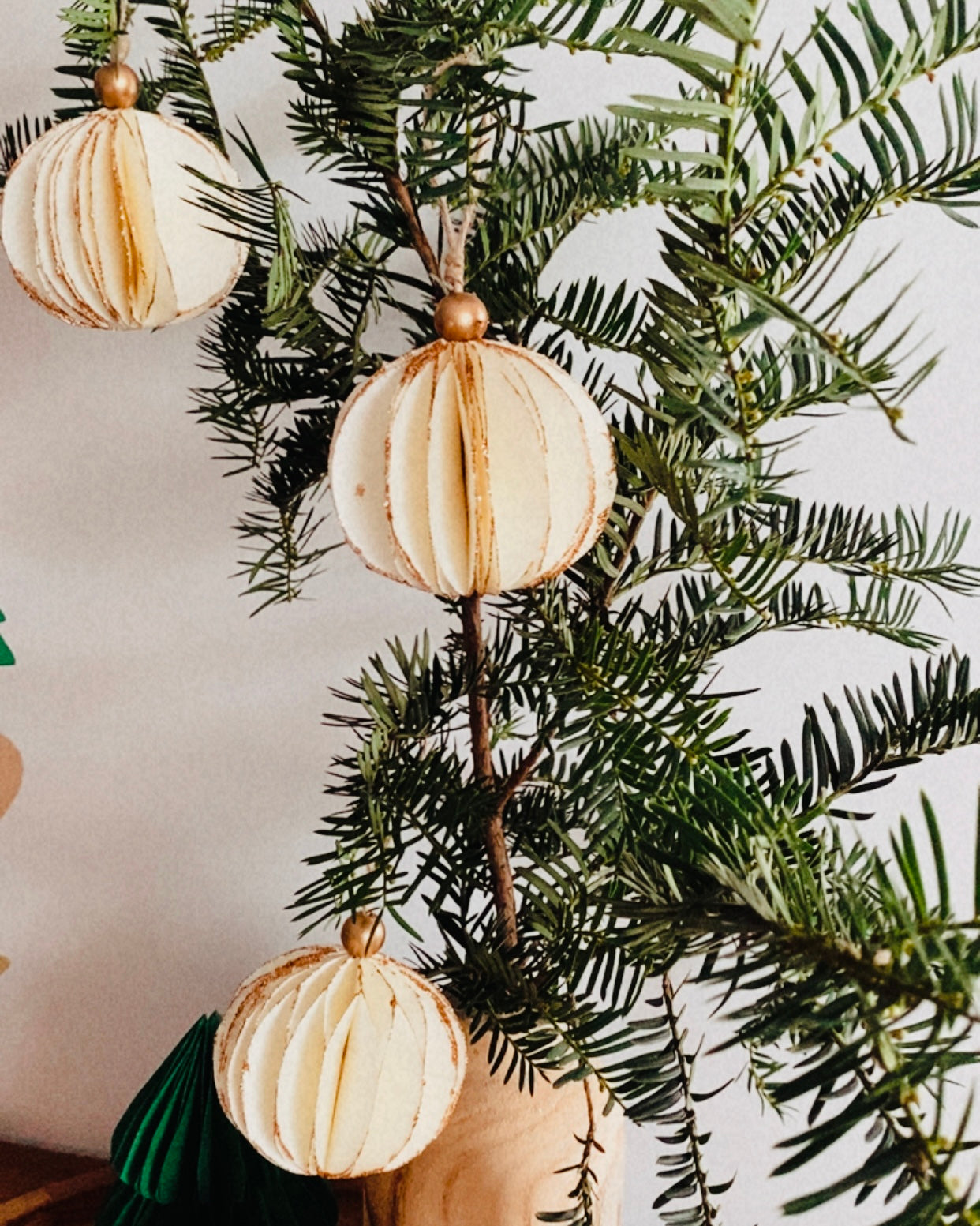 Boules de Noël écru suspendues à une branche de sapin, dévoilant leur forme ronde, légère et texturée. Les paillettes dorées ajoutent une touche scintillante discrète et élégante. L’ensemble crée une ambiance décorative naturelle, apaisante et raffinée, idéale pour une décoration de Noël minimaliste et chaleureuse. ninona
