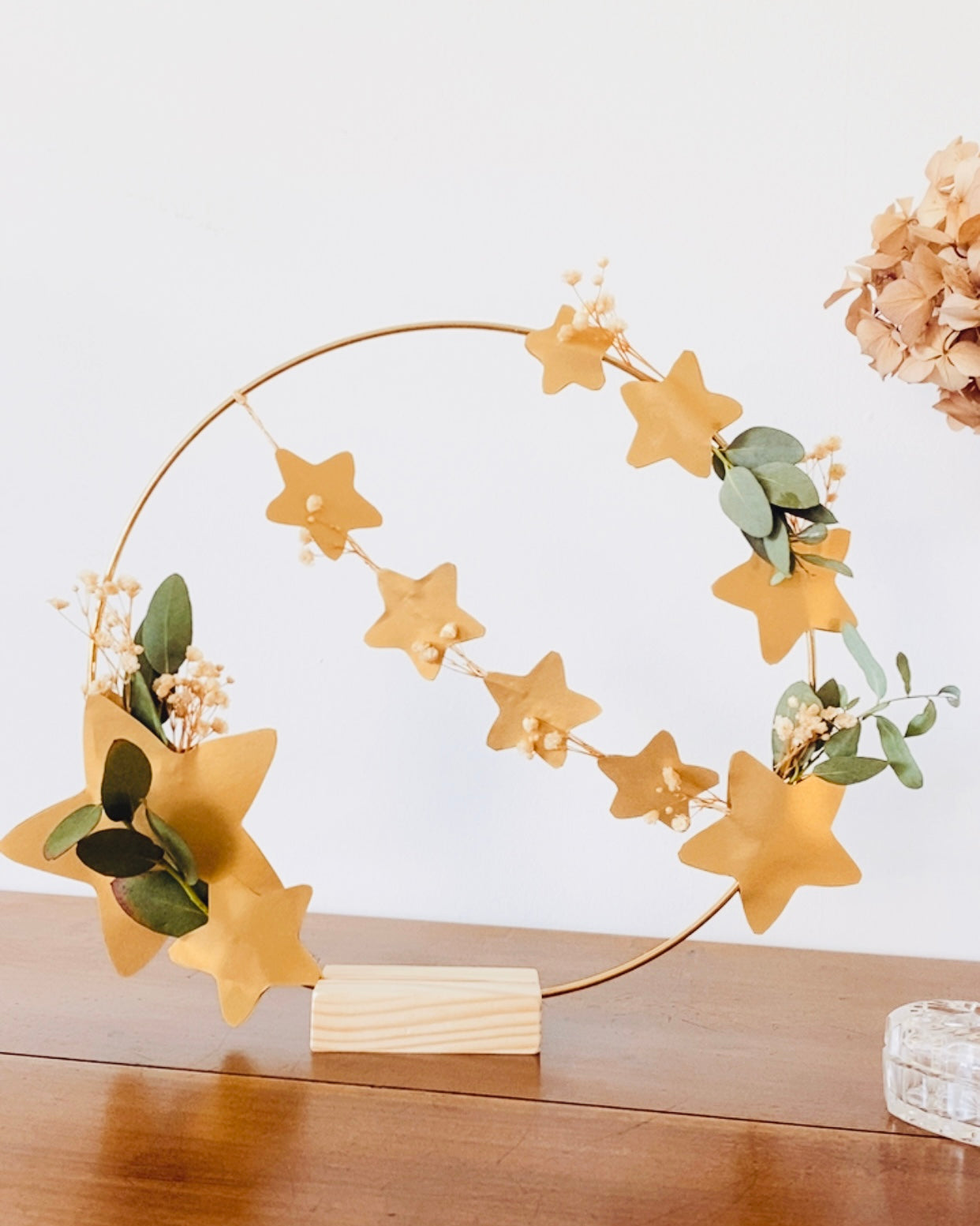 Couronne florale en fleurs séchées et eucalyptus sur anneau doré, décorée d’étoiles dorées en papier découpé. Présentée avec un socle en bois brut pour être exposée sur une étagère, une cheminée ou une table. Création artisanale française, réalisée à la main. Convient pour une déco de Noël chaleureuse ou une décoration intérieure bohème toute l’année. Idéale comme cadeau fait main original et durable. ninona