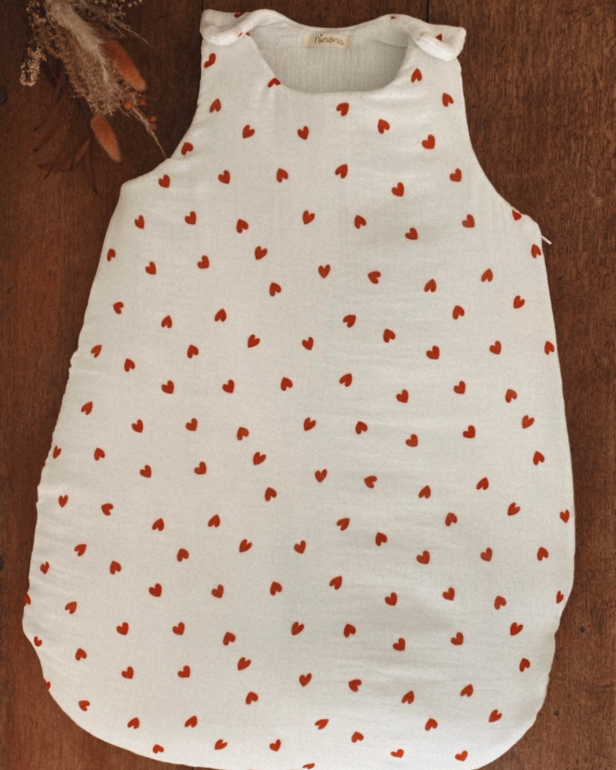 Vue complète d’une gigoteuse bébé hiver en double gaze de coton écrue à petits cœurs rouges. La turbulette est présentée à plat afin de montrer sa forme ample et confortable, pensée pour accompagner les mouvements de bébé pendant son sommeil. Disponible ici en taille 0/6 mois uniquement, elle possède un indice de chaleur TOG 2. Son imprimé cœur apporte une touche tendre et intemporelle, tandis que la double gaze de coton. Une pièce tendre et intemporelle pour compléter le trousseau de naissance.