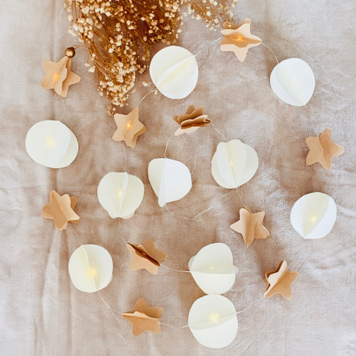 Guirlande lumineuse artisanale composée de délicates formes en papier, mêlant étoiles en kraft et sphères blanches soigneusement pliées à la main. Chaque élément est assemblé sur un fil discret où se glissent de petites LED diffusant une lueur douce et chaleureuse. Cette décoration apporte une ambiance naturelle et poétique, parfaite pour illuminer un intérieur de façon subtile et apaisante. création originale ninona