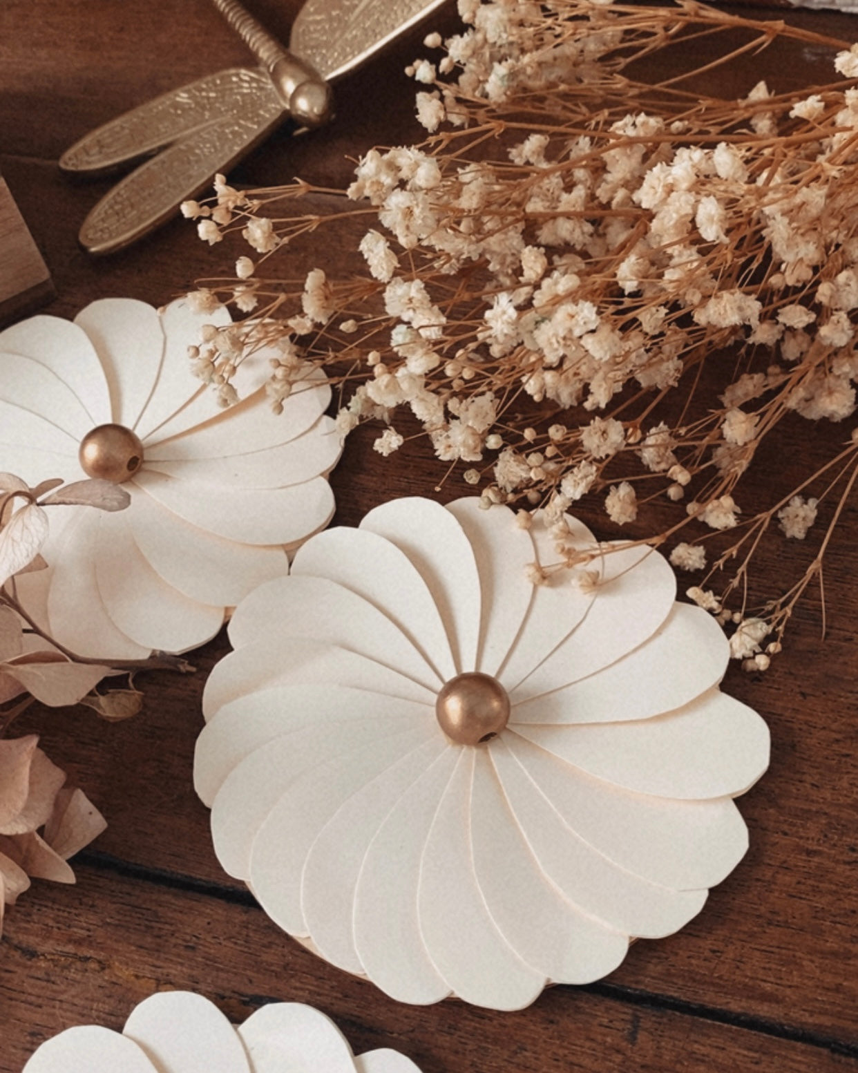 Composition de fleurs en papier écru faites main, photographiées en détail pour mettre en valeur le travail artisanal des pétales et la texture du papier. Les fleurs sont associées à des éléments naturels comme des fleurs séchées et un support en bois, renforçant l’esthétique minimaliste et bohème. Cette décoration florale convient parfaitement à une décoration de table de mariage, un anniversaire ou un intérieur au style naturel et épuré. ninona creation