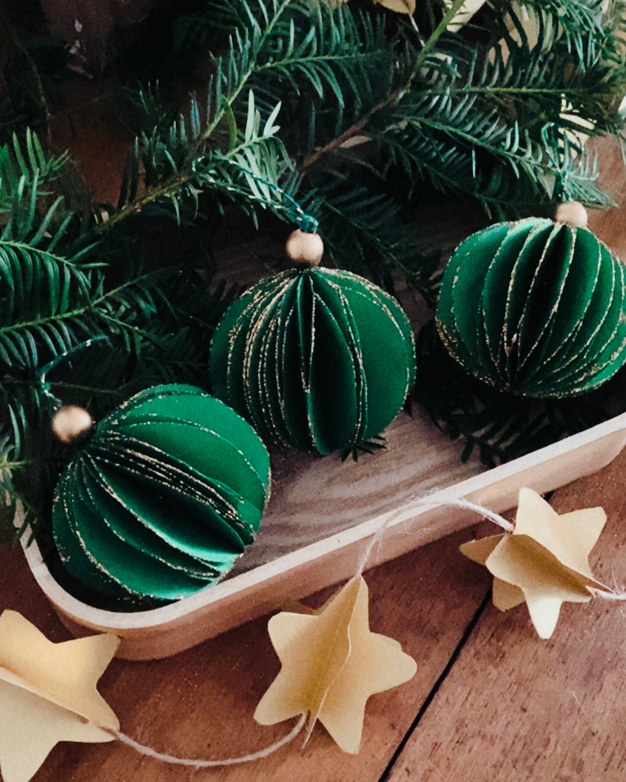 Boule de Noël artisanale en papier vert émeraude, tenue dans une main, montrant les détails délicats du pliage et des fines paillettes dorées. L’ornement dégage une ambiance chaleureuse et naturelle, parfaite pour décorer un sapin ou une branche végétale lors des fêtes de fin d’année. L’image met en valeur le travail fait main et le charme poétique de la décoration, dans une atmosphère douce et authentique. ninona