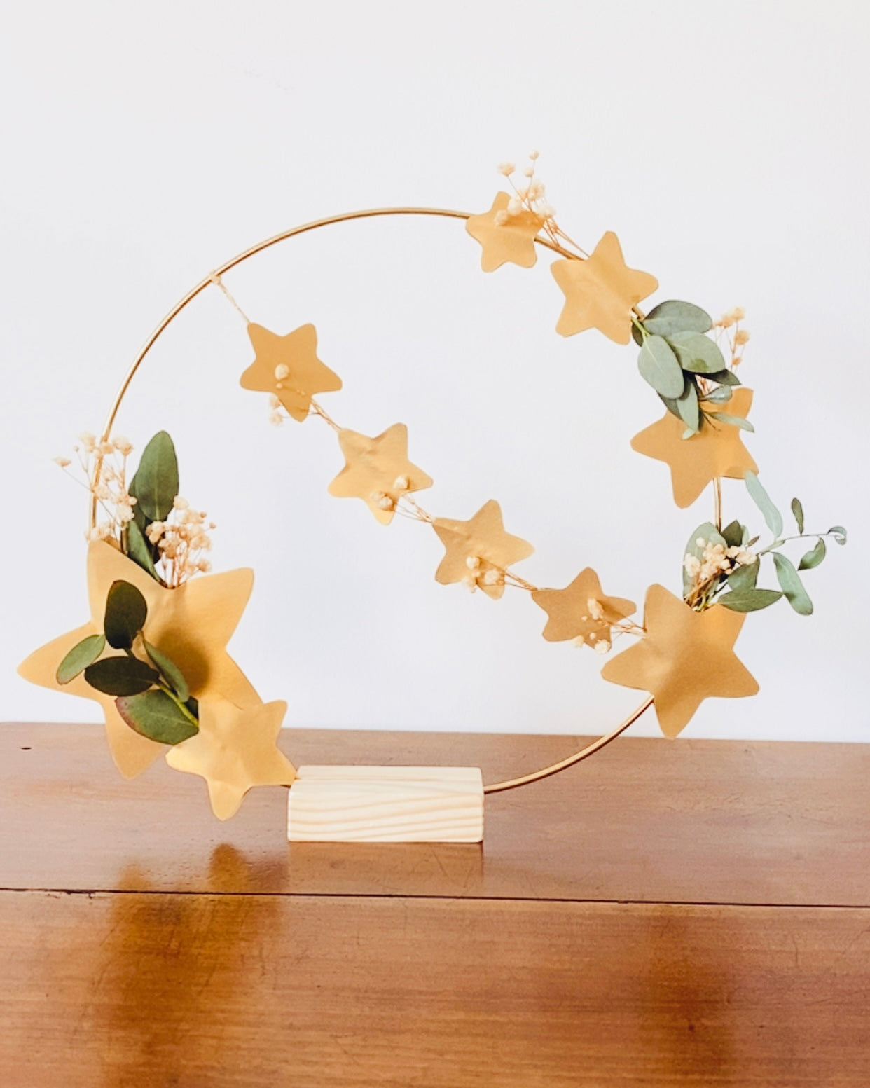 Couronne florale délicate avec eucalyptus et petites fleurs séchées, décorée d’étoiles dorées. Fait main en France, avec soin et attention portée aux détails. Peut être suspendue ou utilisée comme décoration de table avec son socle en bois. Adaptée à la décoration de maison, aux fêtes de fin d’année, à un intérieur bohème ou scandinave. Cadeau chaleureux, durable et symbolique pour Noël ou une occasion spéciale. ninona