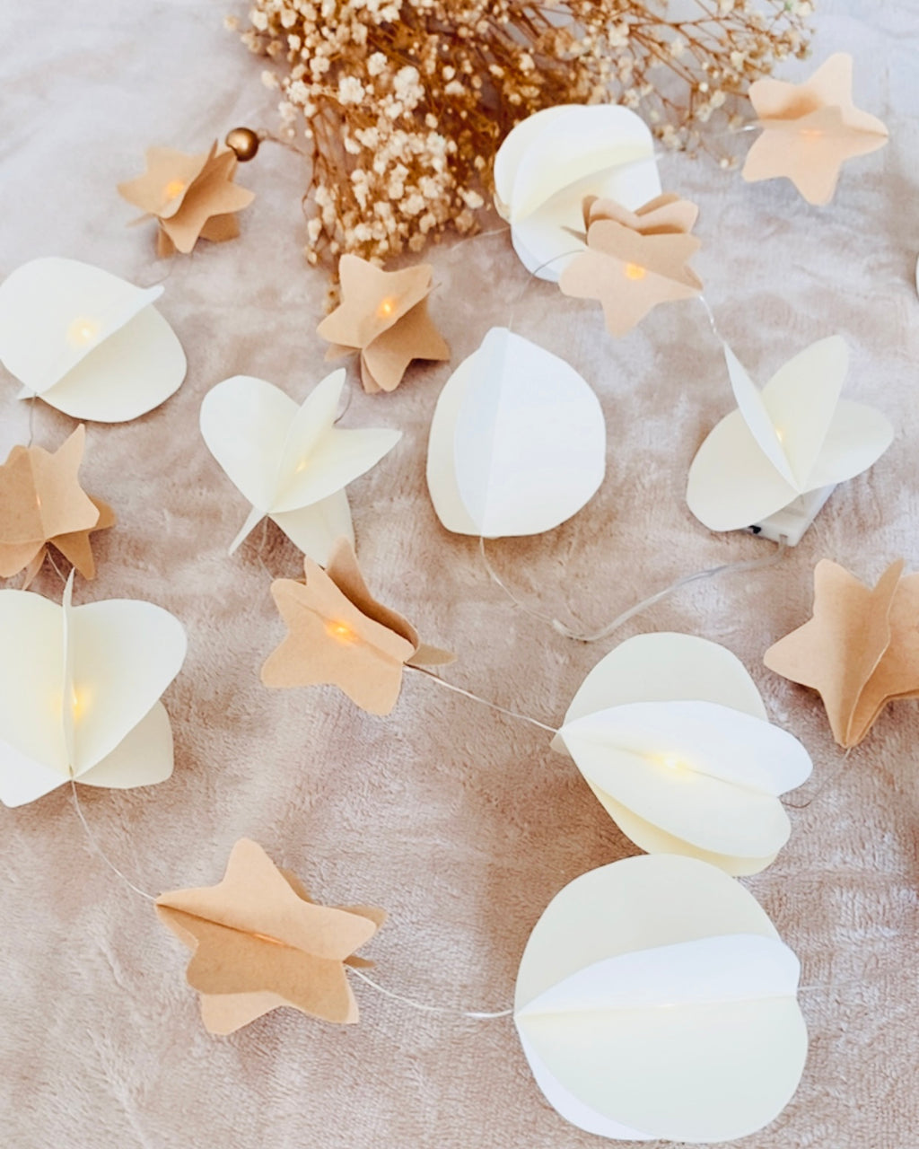 Guirlande décorative réalisée entièrement à la main, composée de petites étoiles en papier brun et de formes rondes blanches montées sur un fil transparent. De discrètes LED diffusent une lumière chaude à travers les volumes en papier, créant un effet doux et délicat. Ce décor artisanal évoque la légèreté, la nature et la poésie, idéal pour embellir une pièce avec une touche authentique. ninona