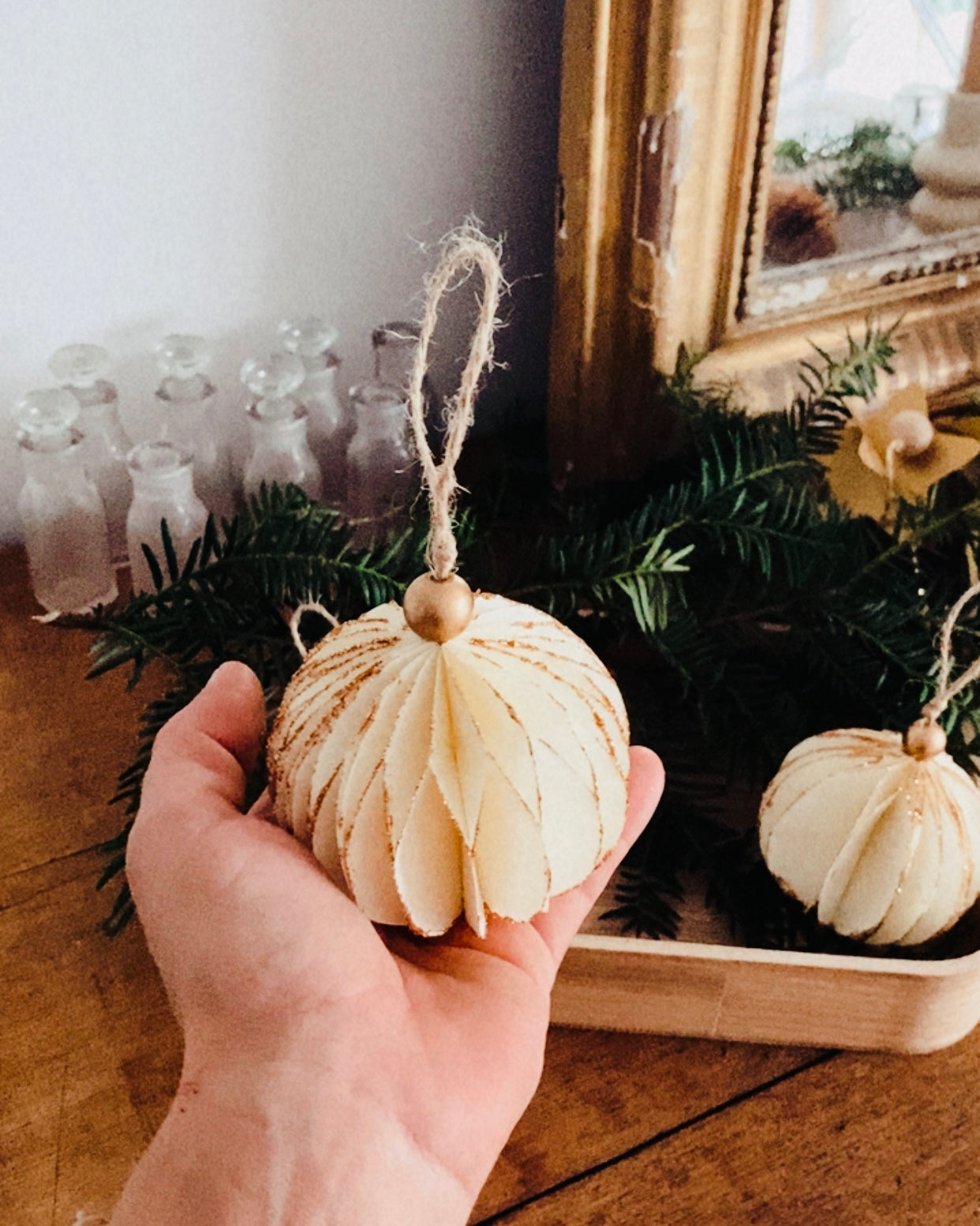 Boules de Noël écru suspendues à une branche de sapin, dévoilant leur forme ronde, légère et texturée. Les paillettes dorées ajoutent une touche scintillante discrète et élégante. L’ensemble crée une ambiance décorative naturelle, apaisante et raffinée, idéale pour une décoration de Noël minimaliste et chaleureuse. ninona