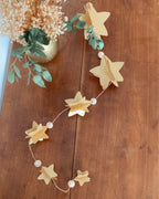 Guirlande murale artisanale en papier avec étoiles 3D et perles en bois, suspendue devant un miroir doré. Cette décoration faite à la main offre une ambiance chaleureuse et lumineuse pour Noël ou pour toute l’année. Sa ficelle dorée apporte une touche festive délicate. Idéale pour salon, chambre ou entrée. Fabrication française soignée, parfaite pour un intérieur inspiré du naturel et du raffinement. ninona