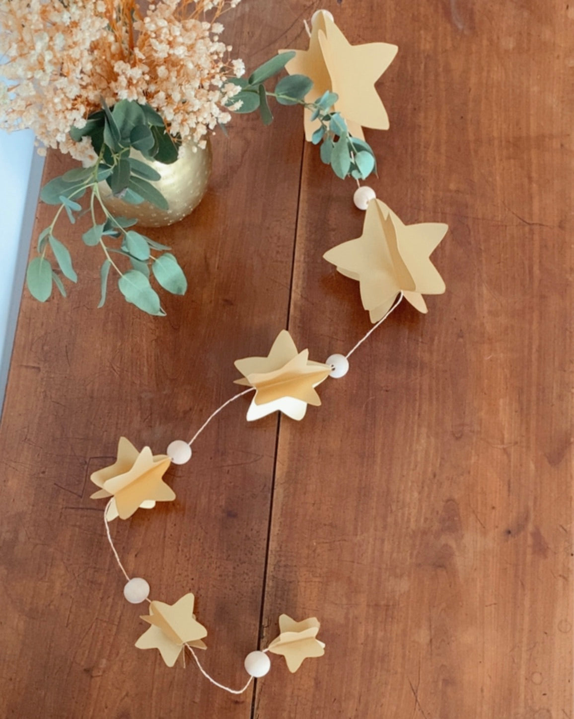 Guirlande murale artisanale en papier avec étoiles 3D et perles en bois, suspendue devant un miroir doré. Cette décoration faite à la main offre une ambiance chaleureuse et lumineuse pour Noël ou pour toute l’année. Sa ficelle dorée apporte une touche festive délicate. Idéale pour salon, chambre ou entrée. Fabrication française soignée, parfaite pour un intérieur inspiré du naturel et du raffinement. ninona