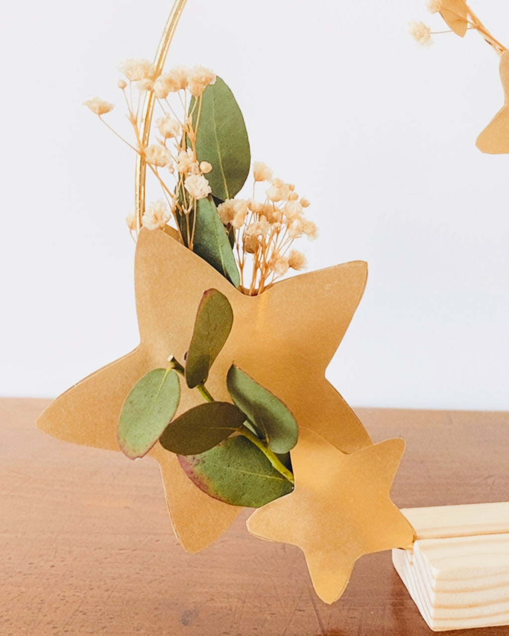 Décoration artisanale élégante : couronne en métal doré ornée de fleurs séchées et de feuilles d’eucalyptus, embellie de petites étoiles dorées. Création faite main en atelier en France. Peut être accrochée en décoration murale ou présentée sur un socle en bois. Parfaite pour une ambiance douce à Noël ou tout au long de l’année. Cadeau idéal pour les amateurs de décoration naturelle et poétique. ninona