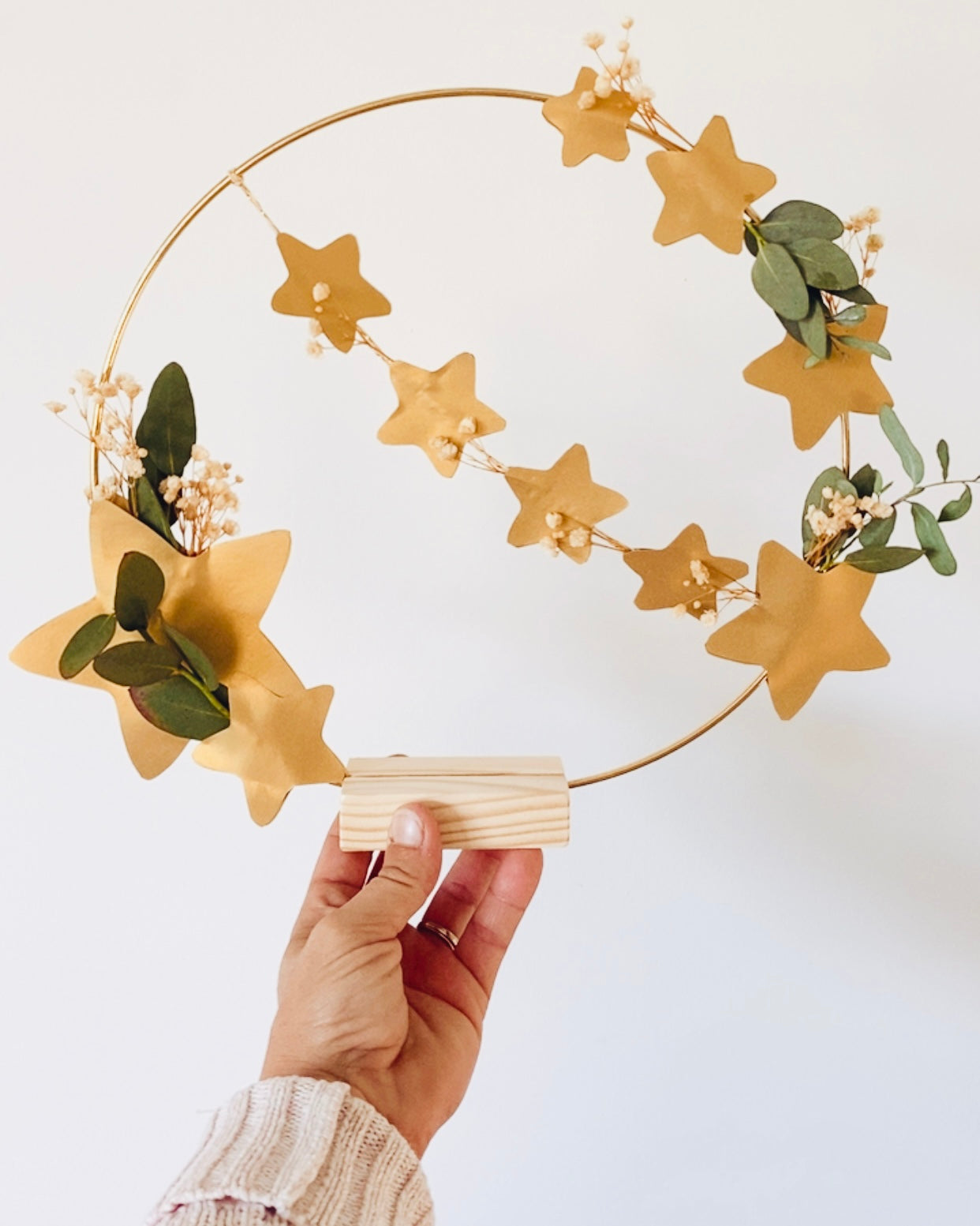 Anneau décoratif doré orné de fleurs séchées et d’eucalyptus, agrémenté d’étoiles dorées découpées à la main. Style naturel et poétique. Création artisanale française pouvant être accrochée ou posée sur un socle. Idéale pour une décoration de Noël douce et minimaliste ou pour sublimer un intérieur toute l’année. Excellent choix de cadeau fait main, durable, attentionné et unique. ninona
