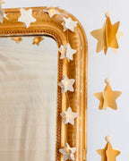 Guirlande décorative faite main en France, composée de six étoiles en papier cartonné assemblées en volume 3D, alternées avec de petites perles en bois naturel et montées sur une ficelle dorée. Décoration légère et poétique, idéale pour Noël, une chambre d’enfant ou pour ajouter une touche chaleureuse à l’intérieur. Chaque étoile est découpée et collée à la main, créant un objet unique et artisanal au style doux et élégant. ninona