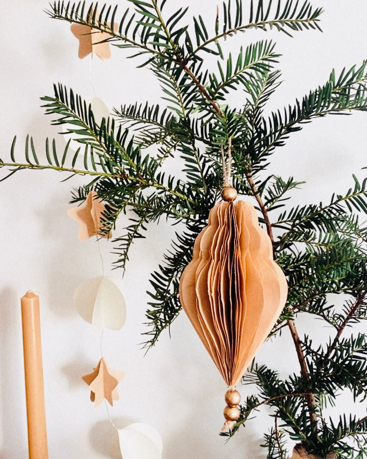 Décoration de Noël artisanale en papier, suspendue par une cordelette naturelle avec une perle dorée. La boule présente une forme élégamment travaillée en plusieurs couches découpées à la main, avec de légères touches de paillettes dorées qui soulignent ses contours. L’ornement, de teinte écru douce, est tenu dans une main, mettant en valeur son relief, sa finesse et son allure poétique, idéale pour décorer un sapin ou une guirlande de fête. ninona