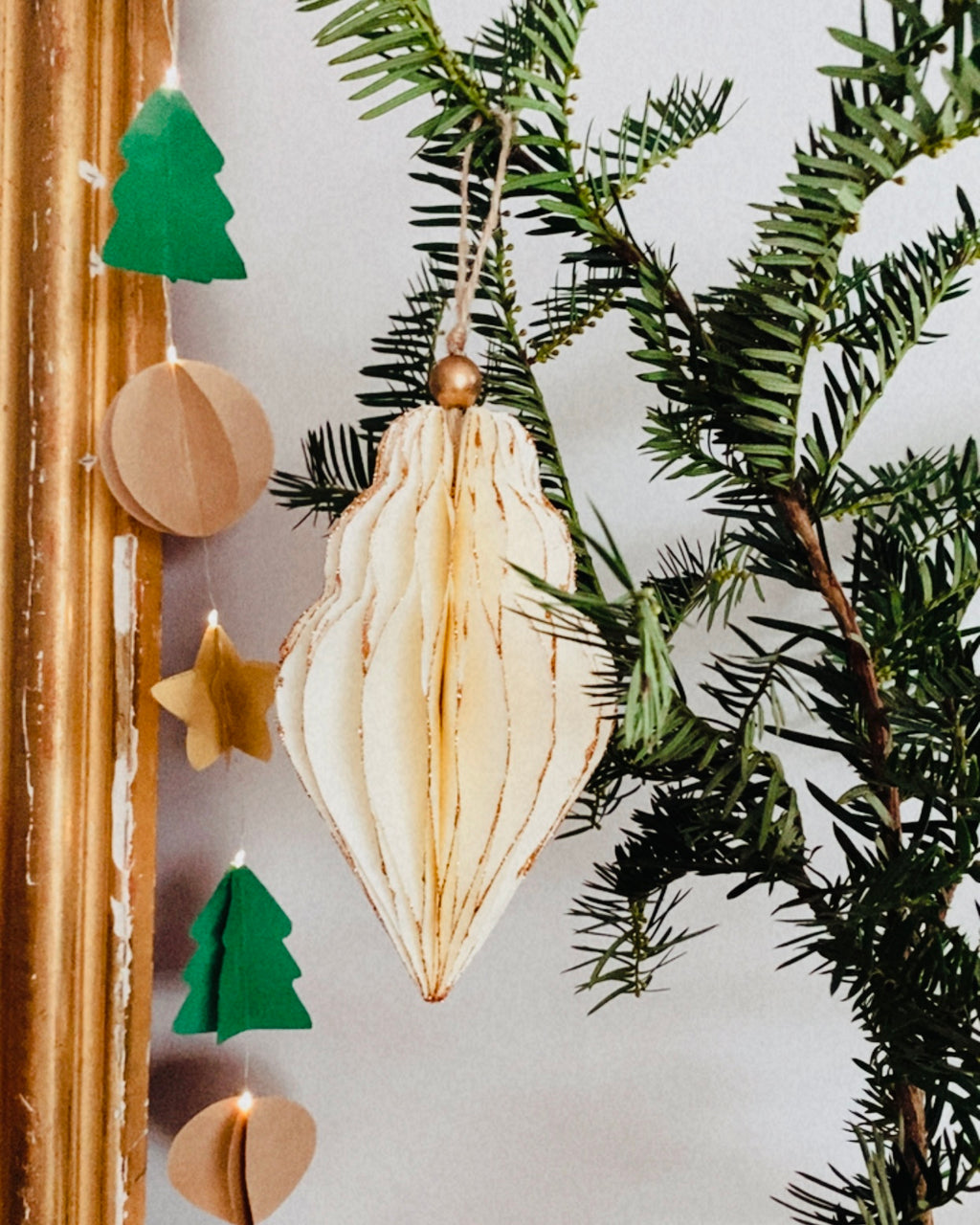 Décoration de Noël artisanale en papier, suspendue par une cordelette naturelle avec une perle dorée. La boule présente une forme élégamment travaillée en plusieurs couches découpées à la main, avec de légères touches de paillettes dorées qui soulignent ses contours. L’ornement, de teinte écru douce, est tenu dans une main, mettant en valeur son relief, sa finesse et son allure poétique, idéale pour décorer un sapin ou une guirlande de fête. ninona