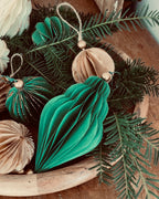 Ornements de Noël faits main en papier, présentés dans une coupe en bois garnie de branches de sapin. Les boules sont de différentes formes et couleurs : vert émeraude profond et écru chaleureux. Certaines sont rehaussées de fines paillettes dorées. L’ensemble évoque une décoration naturelle, authentique et poétique, parfaite pour un Noël doux, artisanal et inspiré par la nature. ninona
