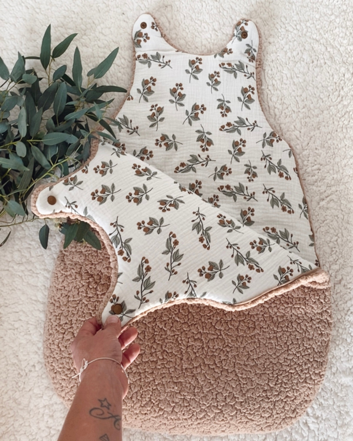 Gigoteuse hiver bébé 0/6 mois ouverte pour révéler son intérieur en double gaze de coton blanc cassé à motifs végétaux. Les petites branches, feuilles et baies dans des tons bruns et verts contrastent délicatement avec le sherpa chocolat. Cette turbulette TOG 2 est pensée comme un véritable cocon chaud et douillet pour les nuits d’hiver. Confectionnée artisanalement avec soin, elle associe confort, douceur, charme naturel et esprit bohème pour la chambre de bébé. Une pièce bébé, douce et belle. Ninona