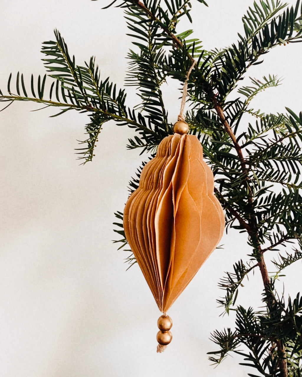 Décoration de Noël artisanale en papier, suspendue par une cordelette naturelle avec une perle dorée. La boule présente une forme élégamment travaillée en plusieurs couches découpées à la main, avec de légères touches de paillettes dorées qui soulignent ses contours. L’ornement, de teinte écru douce, est tenu dans une main, mettant en valeur son relief, sa finesse et son allure poétique, idéale pour décorer un sapin ou une guirlande de fête. ninona
