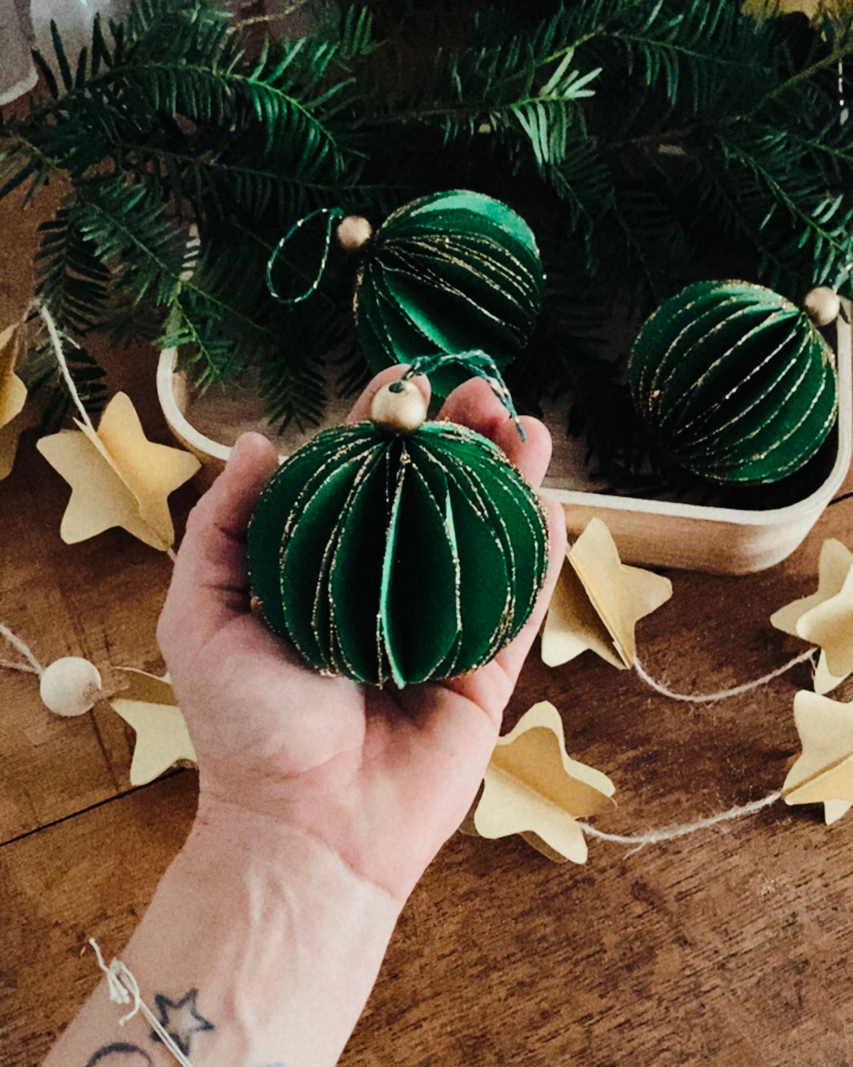 Composition festive réunissant boules de Noël artisanales, plateau en bois et branches de sapin naturel. Le décor évoque la simplicité d’un Noël fait maison, avec des matières naturelles, du papier, du bois et de fines touches dorées. L’ensemble transmet une ambiance calme, chaleureuse et poétique. ninona
