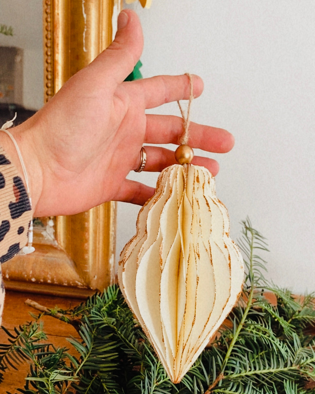 Décoration de Noël artisanale en papier, suspendue par une cordelette naturelle avec une perle dorée. La boule présente une forme élégamment travaillée en plusieurs couches découpées à la main, avec de légères touches de paillettes dorées qui soulignent ses contours. L’ornement, de teinte écru douce, est tenu dans une main, mettant en valeur son relief, sa finesse et son allure poétique, idéale pour décorer un sapin ou une guirlande de fête. ninona