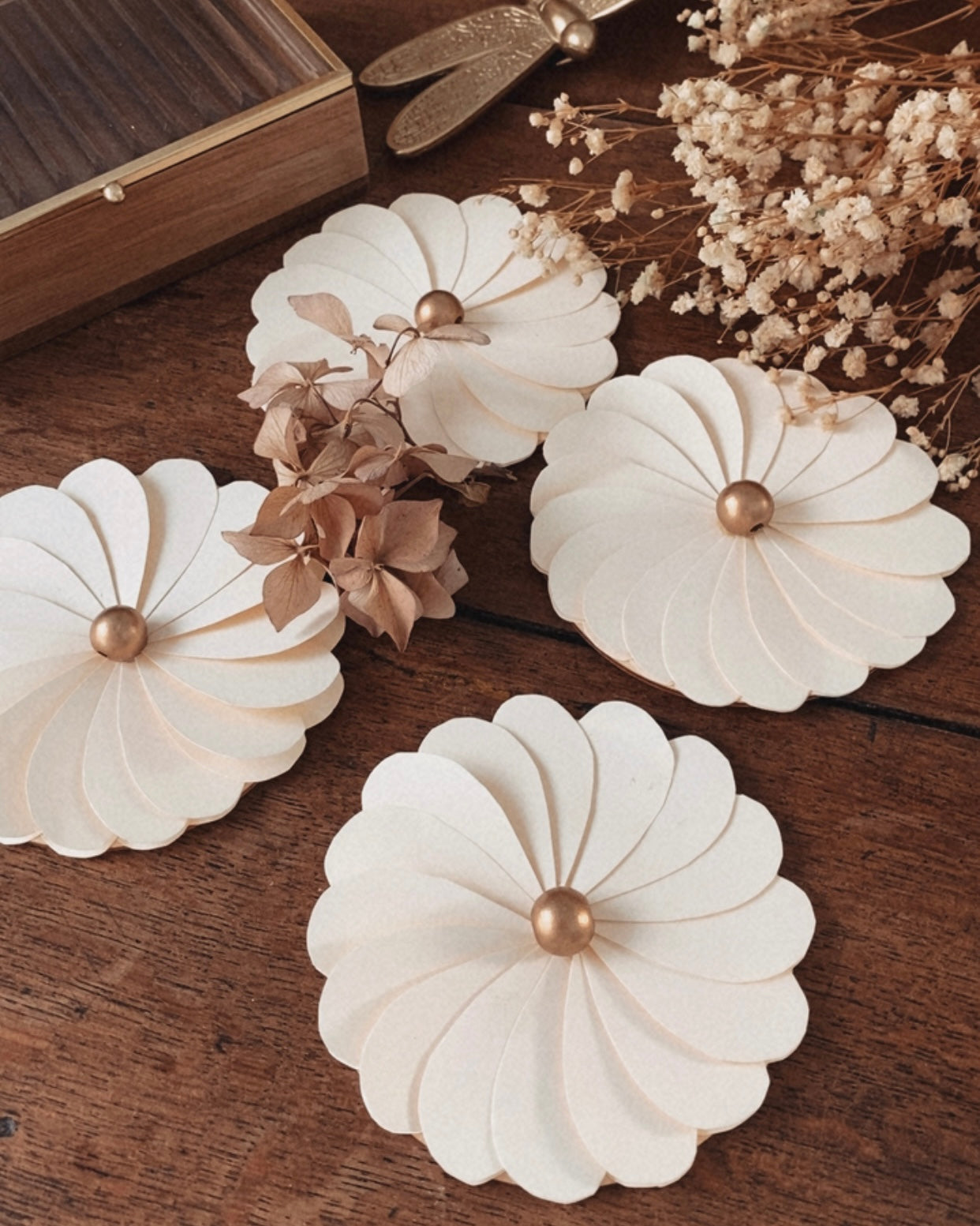 Composition de fleurs en papier écru faites main, photographiées en détail pour mettre en valeur le travail artisanal des pétales et la texture du papier. Les fleurs sont associées à des éléments naturels comme des fleurs séchées et un support en bois, renforçant l’esthétique minimaliste et bohème. Cette décoration florale convient parfaitement à une décoration de table de mariage, un anniversaire ou un intérieur au style naturel et épuré. ninona creation