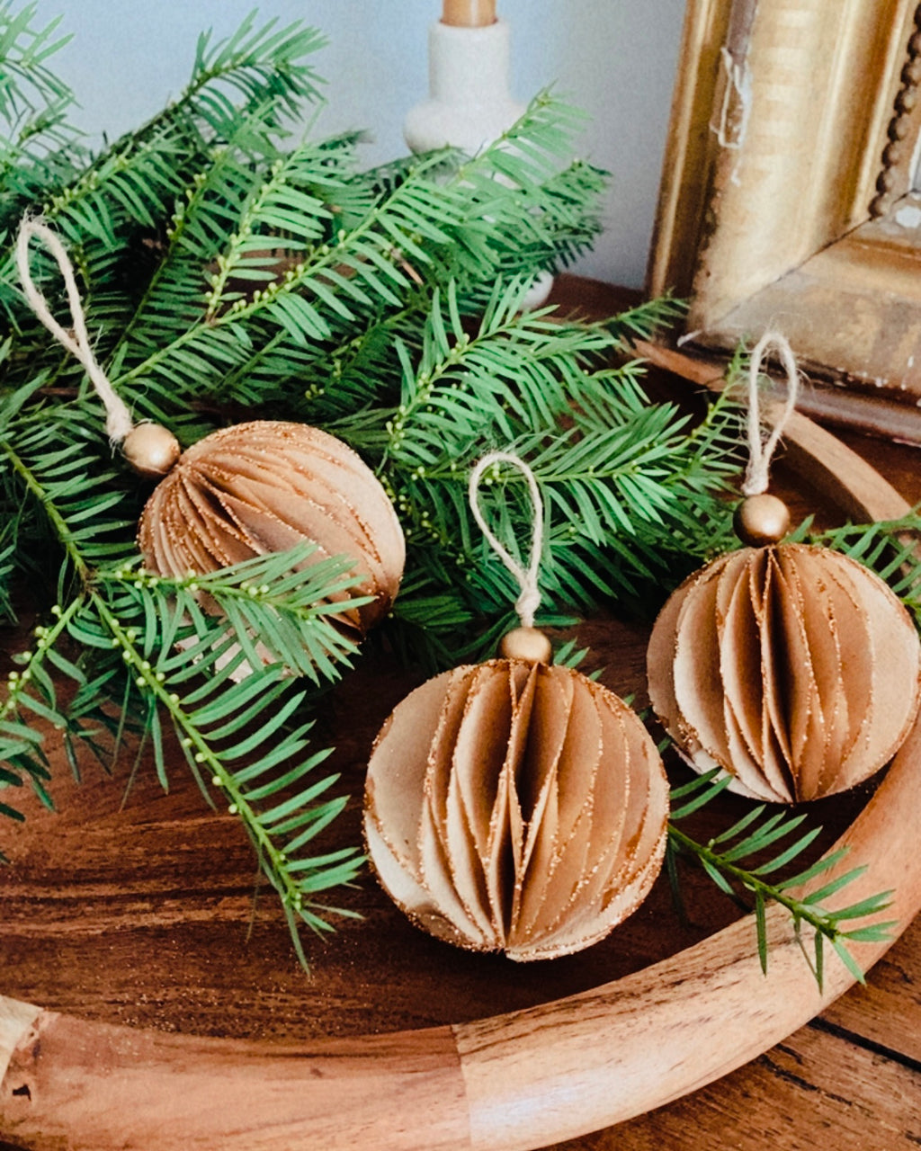 Lot de trois boules de Noël marron posées dans un plateau ou sur une table en bois, entourées de branches de sapin naturelles. L’atmosphère est douce, rustique et chaleureuse, évoquant une décoration de fêtes naturelle et artisanale. Les paillettes apportent une touche délicate de lumière et de magie. ninona
