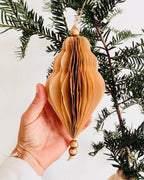 Décoration de Noël artisanale en papier, suspendue par une cordelette naturelle avec une perle dorée. La boule présente une forme élégamment travaillée en plusieurs couches découpées à la main, avec de légères touches de paillettes dorées qui soulignent ses contours. L’ornement, de teinte écru douce, est tenu dans une main, mettant en valeur son relief, sa finesse et son allure poétique, idéale pour décorer un sapin ou une guirlande de fête. ninona