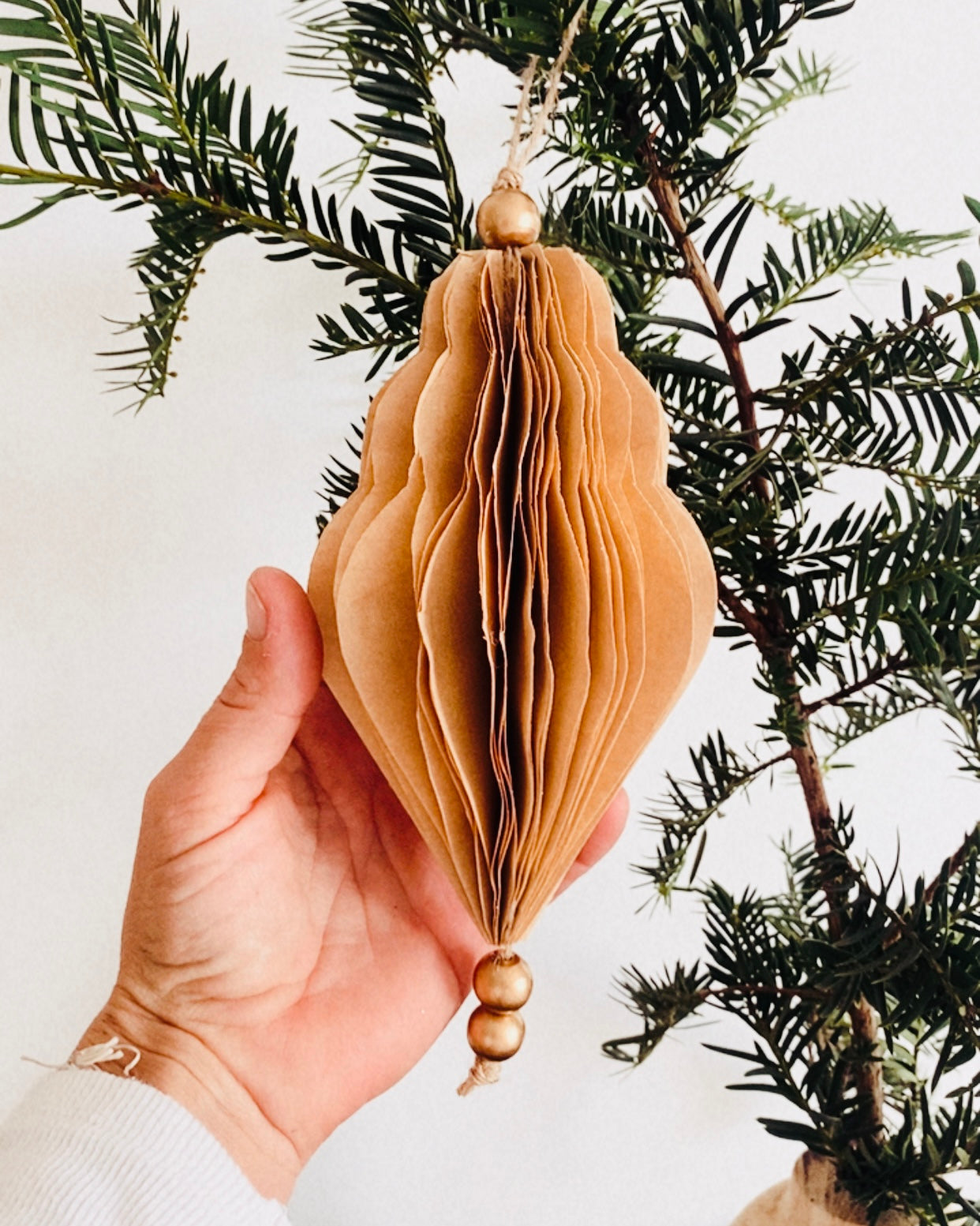 Décoration de Noël artisanale en papier, suspendue par une cordelette naturelle avec une perle dorée. La boule présente une forme élégamment travaillée en plusieurs couches découpées à la main, avec de légères touches de paillettes dorées qui soulignent ses contours. L’ornement, de teinte écru douce, est tenu dans une main, mettant en valeur son relief, sa finesse et son allure poétique, idéale pour décorer un sapin ou une guirlande de fête. ninona