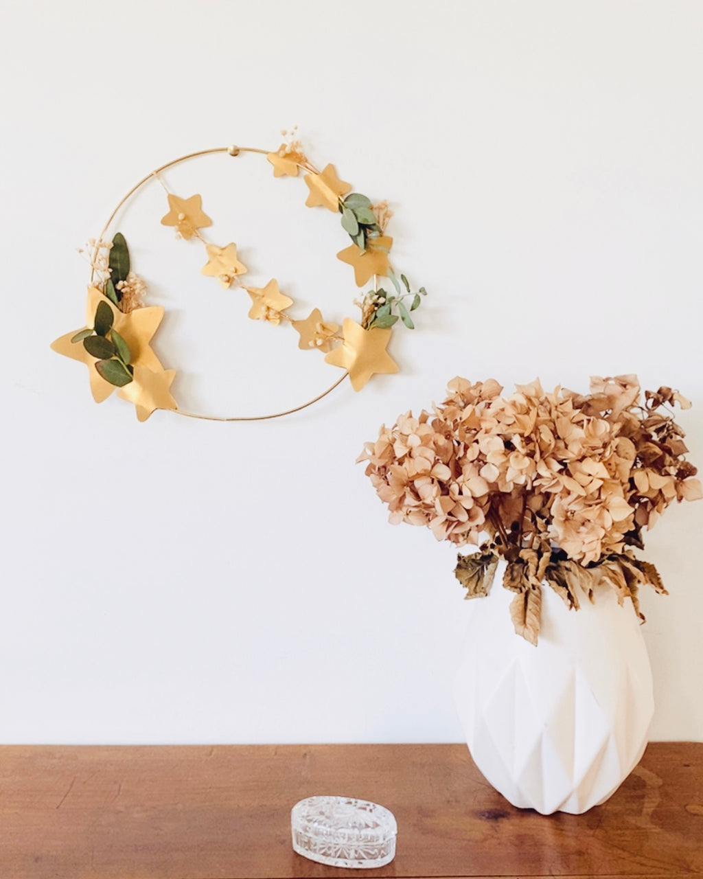 Couronne décorative minimaliste, composée d’un cercle en métal doré, de fleurs séchées et d’eucalyptus, avec une guirlande d’étoiles dorées. Réalisée à la main en France. Peut être placée sur un socle en bois ou accrochée au mur. Parfaite pour un décor de Noël naturel ou une touche délicate dans une chambre ou un salon. Cadeau artisanal original pour les fêtes ou pour offrir une décoration intemporelle. ninona