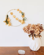 Couronne décorative minimaliste, composée d’un cercle en métal doré, de fleurs séchées et d’eucalyptus, avec une guirlande d’étoiles dorées. Réalisée à la main en France. Peut être placée sur un socle en bois ou accrochée au mur. Parfaite pour un décor de Noël naturel ou une touche délicate dans une chambre ou un salon. Cadeau artisanal original pour les fêtes ou pour offrir une décoration intemporelle. ninona