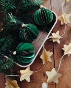 Boule de Noël artisanale en papier vert émeraude, tenue dans une main, montrant les détails délicats du pliage et des fines paillettes dorées. L’ornement dégage une ambiance chaleureuse et naturelle, parfaite pour décorer un sapin ou une branche végétale lors des fêtes de fin d’année. L’image met en valeur le travail fait main et le charme poétique de la décoration, dans une atmosphère douce et authentique. ninona