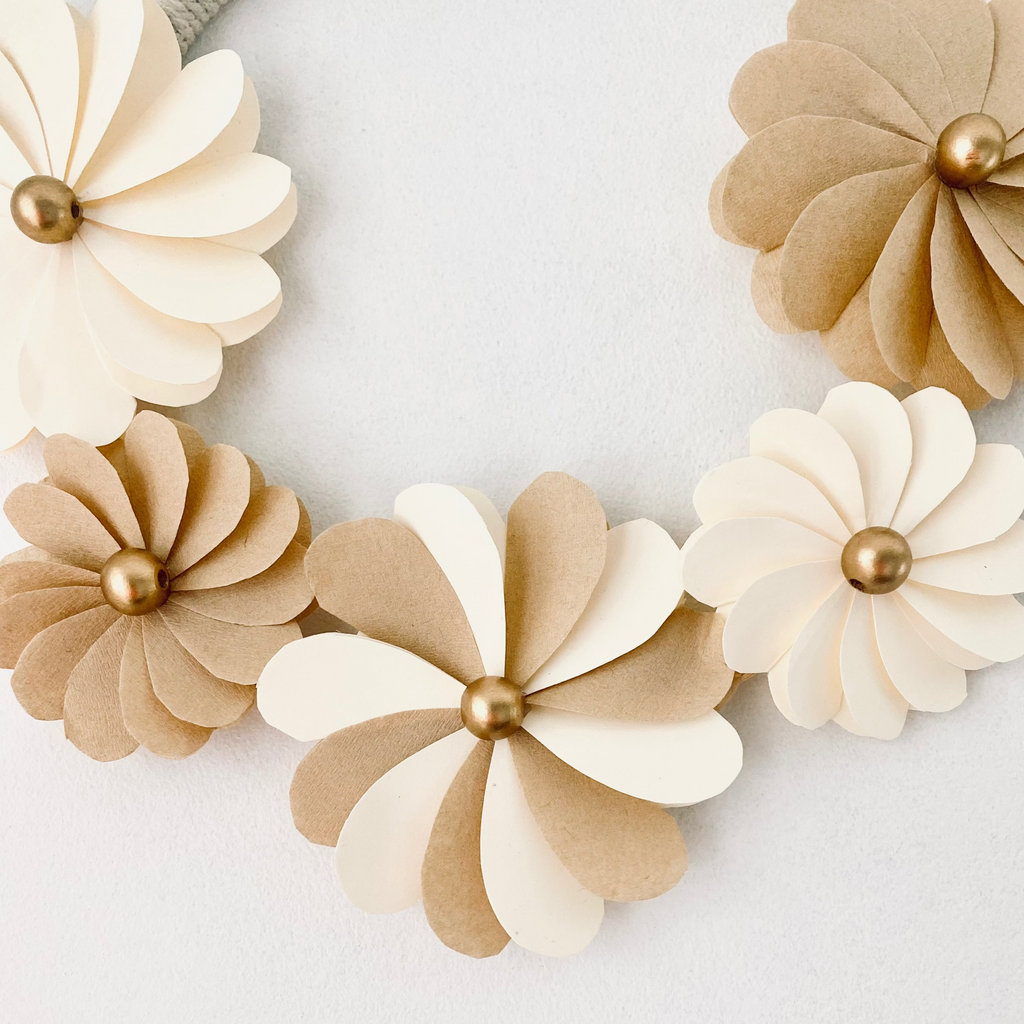Décoration murale en forme de couronne florale, entièrement créée à la main. Les fleurs sont fabriquées avec des pétales en papier écru et filtres à café, soigneusement découpés et collés. Chaque fleur est embellie d’une perle de bois cuivrée au centre. Parfaite pour un intérieur chaleureux, une ambiance bohème ou un cadeau artisanal. Couronne légère et poétique, idéale pour sublimer toute pièce. ninona