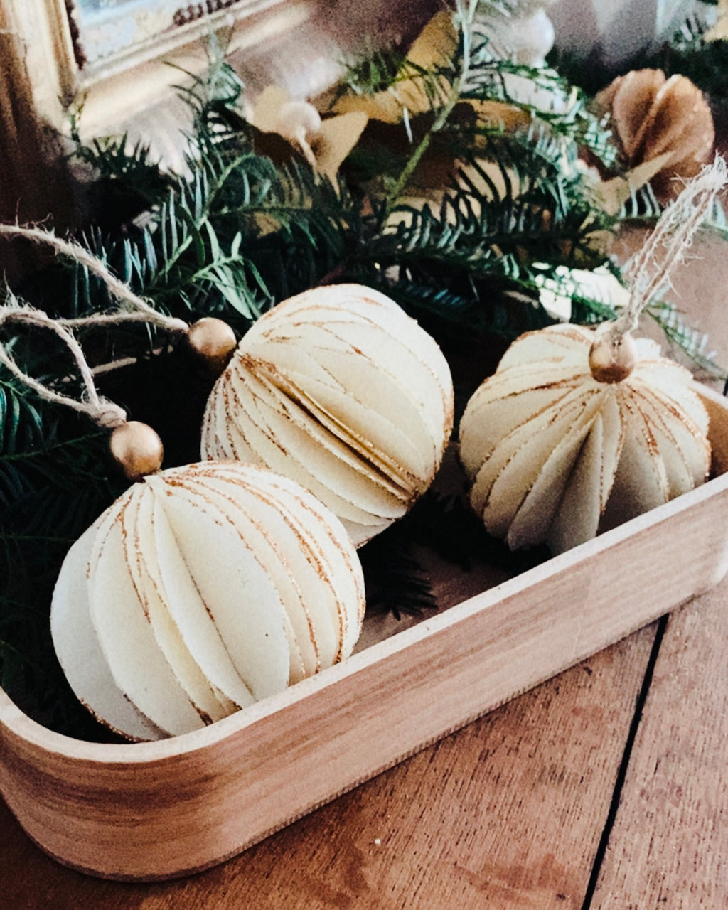 Trois boules de Noël artisanales écru reposant dans un petit plateau en bois entouré de branches de sapin naturel. Les paillettes dorées soulignent les courbes du papier, créant une ambiance douce, lumineuse et poétique. La scène évoque la chaleur d’un Noël artisanal, simple et authentique, où les matières naturelles sont mises à l’honneur. ninona