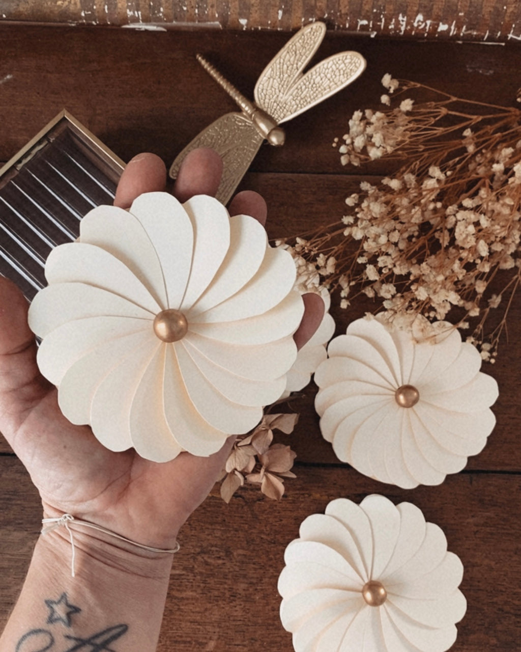Fleur en papier écru tenue dans la main, permettant d’apprécier son diamètre, son volume et la finesse de sa fabrication artisanale. Les pétales découpés avec précision forment une fleur délicate, rehaussée d’une perle en bois au centre. Cette décoration en papier est idéale comme centre de table, élément décoratif mural ou accessoire de décoration pour un mariage, une fête ou une chambre d’enfant au style doux et naturel. ninona creation