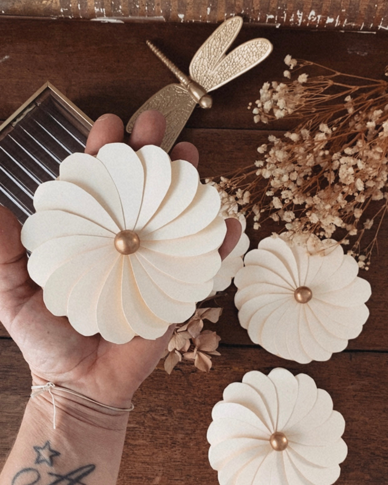 Fleur en papier écru tenue dans la main, permettant d’apprécier son diamètre, son volume et la finesse de sa fabrication artisanale. Les pétales découpés avec précision forment une fleur délicate, rehaussée d’une perle en bois au centre. Cette décoration en papier est idéale comme centre de table, élément décoratif mural ou accessoire de décoration pour un mariage, une fête ou une chambre d’enfant au style doux et naturel. ninona creation