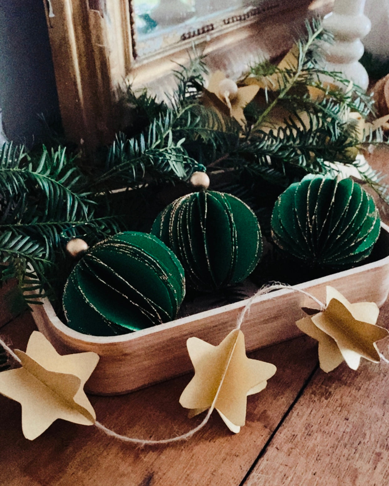 Boule de Noël artisanale en papier vert émeraude, tenue dans une main, montrant les détails délicats du pliage et des fines paillettes dorées. L’ornement dégage une ambiance chaleureuse et naturelle, parfaite pour décorer un sapin ou une branche végétale lors des fêtes de fin d’année. L’image met en valeur le travail fait main et le charme poétique de la décoration, dans une atmosphère douce et authentique. ninona