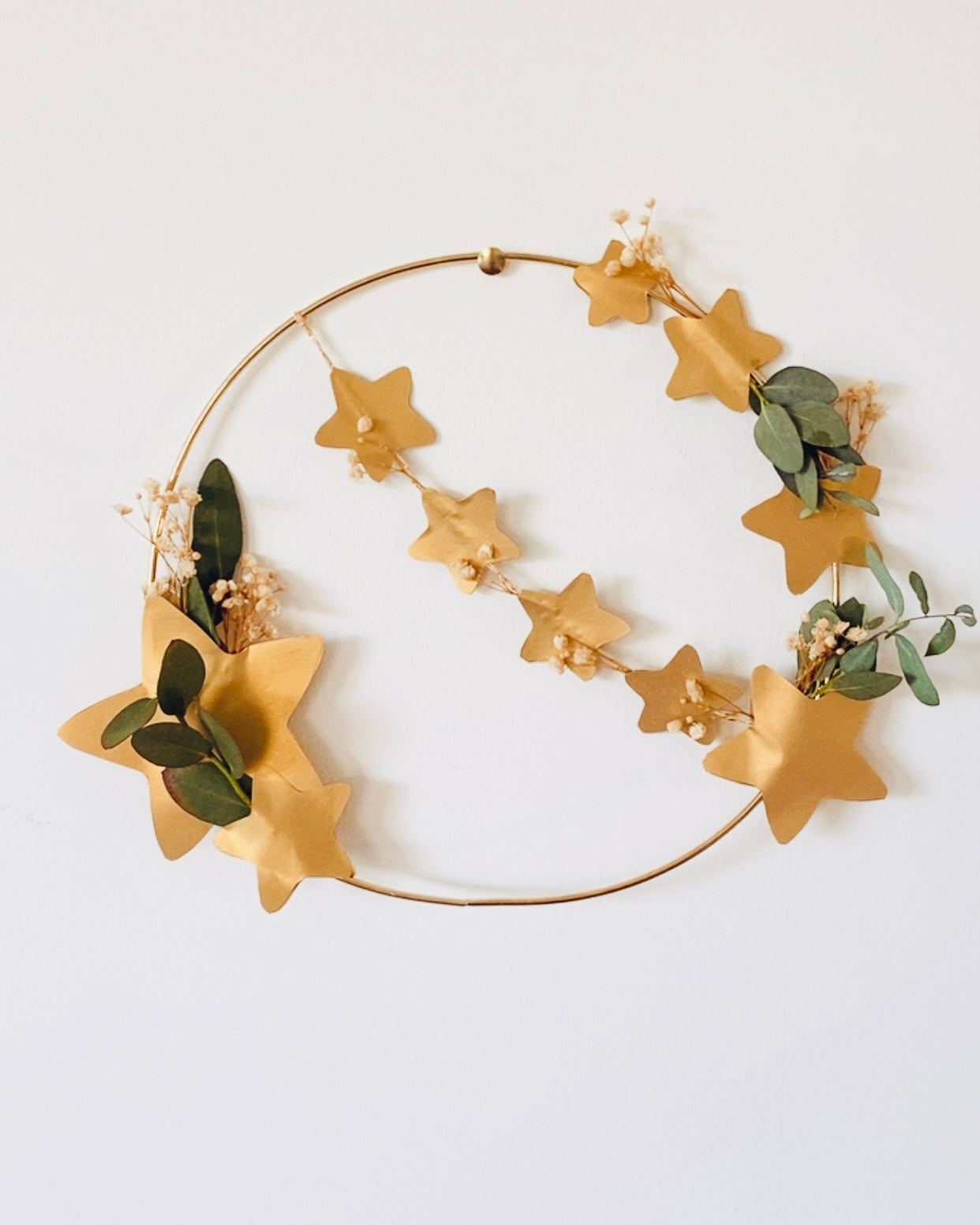 Couronne faite main en atelier français, associant eucalyptus, petites fleurs séchées blanches et étoiles dorées sur un cercle en métal. Présentée avec socle en bois. Décoration idéale pour Noël, une table de fête ou une décoration intérieure douce et naturelle. Convient également toute l’année pour créer une ambiance chaleureuse et apaisante. Cadeau authentique et unique à offrir ou à s’offrir. ninona