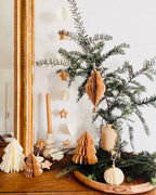 Décoration de Noël artisanale en papier, suspendue par une cordelette naturelle avec une perle dorée. La boule présente une forme élégamment travaillée en plusieurs couches découpées à la main, avec de légères touches de paillettes dorées qui soulignent ses contours. L’ornement, de teinte écru douce, est tenu dans une main, mettant en valeur son relief, sa finesse et son allure poétique, idéale pour décorer un sapin ou une guirlande de fête. ninona