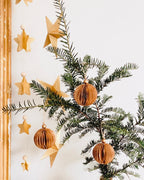 Boules de Noël marron suspendues à une branche dans un intérieur lumineux et épuré. Le contraste entre le vert profond du sapin et la douceur chaleureuse du marron crée une harmonie naturelle. Les paillettes dorées apportent un éclat subtil qui rappelle la magie discrète des fêtes. ninona