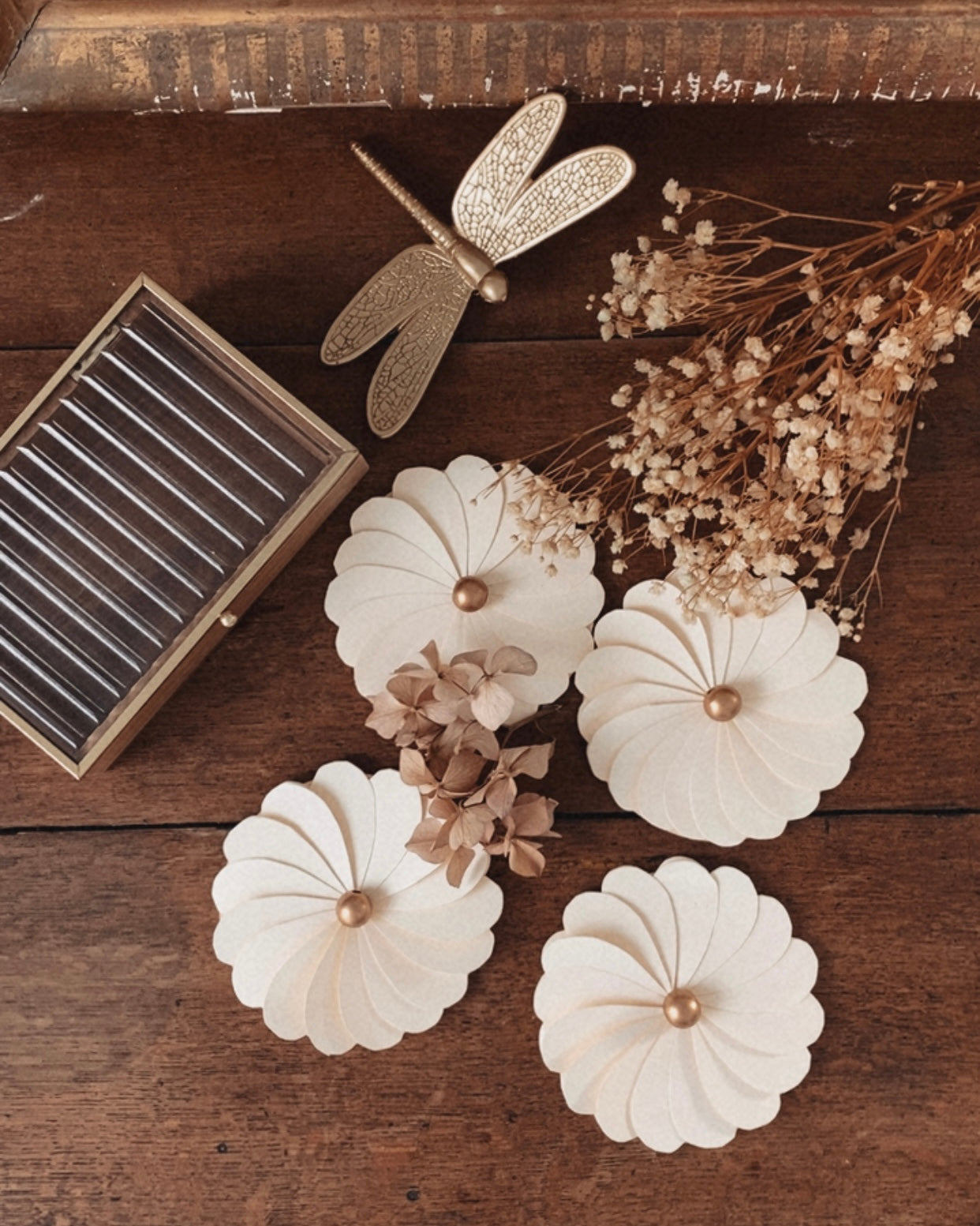 Fleurs décoratives en papier écru, réalisées à la main, disposées sur une surface en bois ancien aux tons chaleureux. Chaque fleur est composée de pétales finement découpés et superposés, avec un cœur en perle de bois. L’ensemble évoque une décoration naturelle et bohème, idéale pour une table de mariage, un centre de table élégant ou une décoration murale artisanale. Ces fleurs en papier apportent une atmosphère douce et poétique à une décoration de fête ou d’intérieur. ninona creation