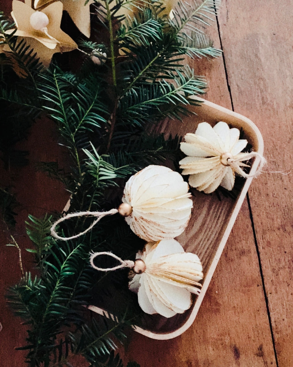 Trois boules de Noël artisanales écru reposant dans un petit plateau en bois entouré de branches de sapin naturel. Les paillettes dorées soulignent les courbes du papier, créant une ambiance douce, lumineuse et poétique. La scène évoque la chaleur d’un Noël artisanal, simple et authentique, où les matières naturelles sont mises à l’honneur. ninona