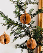 Boules de Noël marron suspendues à une branche dans un intérieur lumineux et épuré. Le contraste entre le vert profond du sapin et la douceur chaleureuse du marron crée une harmonie naturelle. Les paillettes dorées apportent un éclat subtil qui rappelle la magie discrète des fêtes. ninona