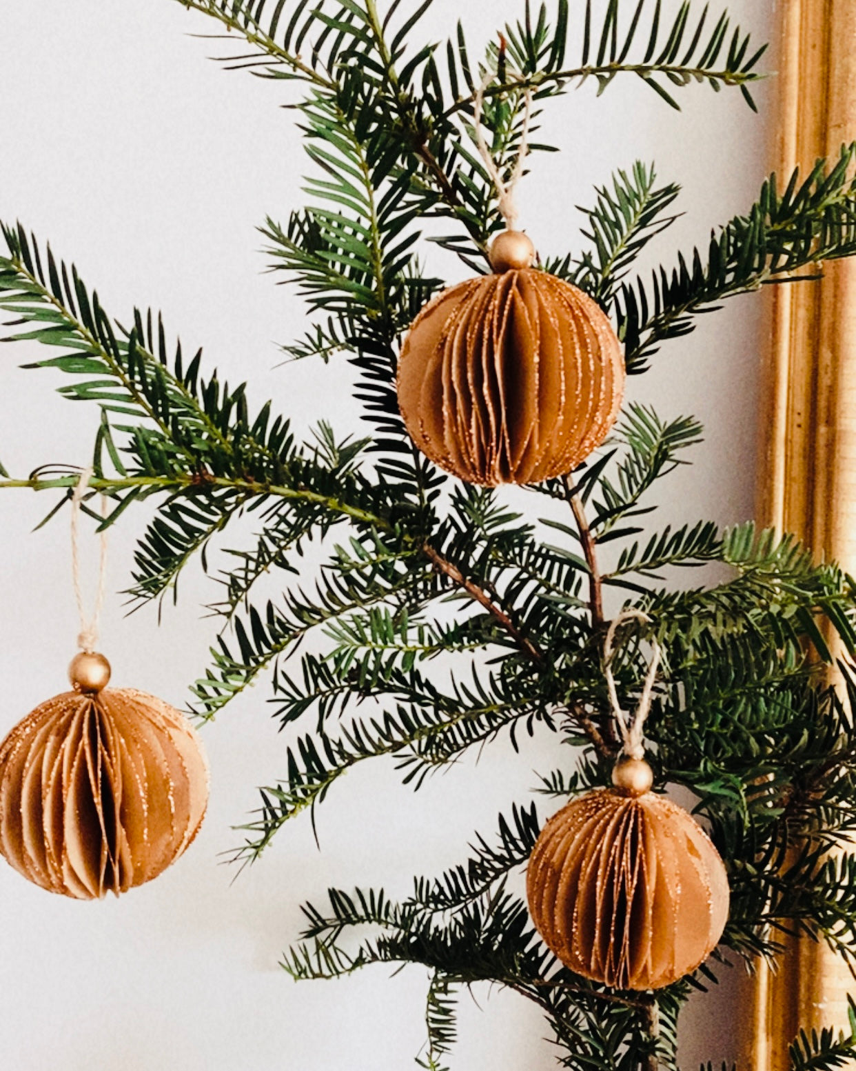 Boules de Noël marron suspendues à une branche dans un intérieur lumineux et épuré. Le contraste entre le vert profond du sapin et la douceur chaleureuse du marron crée une harmonie naturelle. Les paillettes dorées apportent un éclat subtil qui rappelle la magie discrète des fêtes. ninona