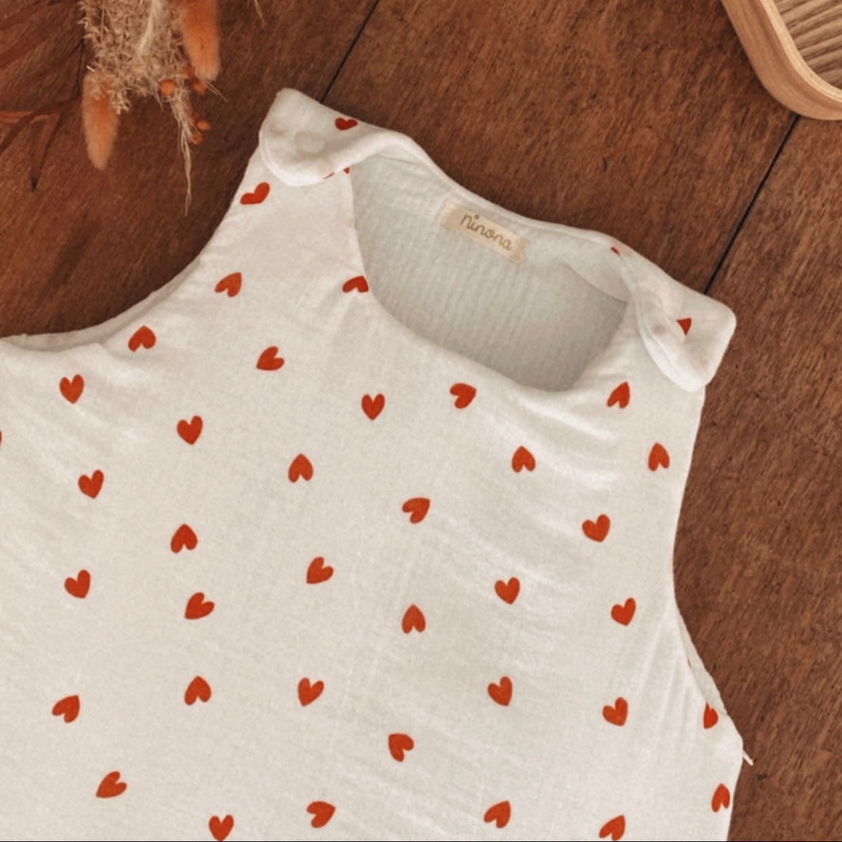 Zoom sur le haut d’une gigoteuse bébé écrue en double gaze de coton, décorée de petits cœurs rouges. On distingue le col arrondi, les emmanchures et les deux pressions placées aux épaules pour faciliter l’habillage. La matière est douce et légèrement gaufrée, avec un intérieur en double gaze de coton. Cette turbulette d’hiver TOG 2 convient aux bébés de 0 à 6 mois. Un modèle délicat et intemporel, idéal pour les nuits fraîches. Une finition délicate qui souligne le soin apporté à chaque détail ! 