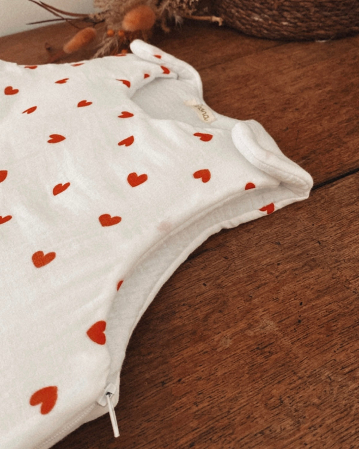 Vue rapprochée de la gigoteuse bébé écrue à petits cœurs rouges, mettant en valeur la texture souple et légèrement gaufrée de la double gaze de coton. On aperçoit la fermeture éclair latérale ainsi que les finitions du bas arrondi. Ce modèle est une gigoteuse d’hiver TOG 2, disponible en taille 0/6 mois. Conçue pour offrir douceur et confort pendant les nuits fraîches, elle se distingue par son motif de petits cœurs. Une ouverture pratique pour installer bébé facilement et préserver son confort.