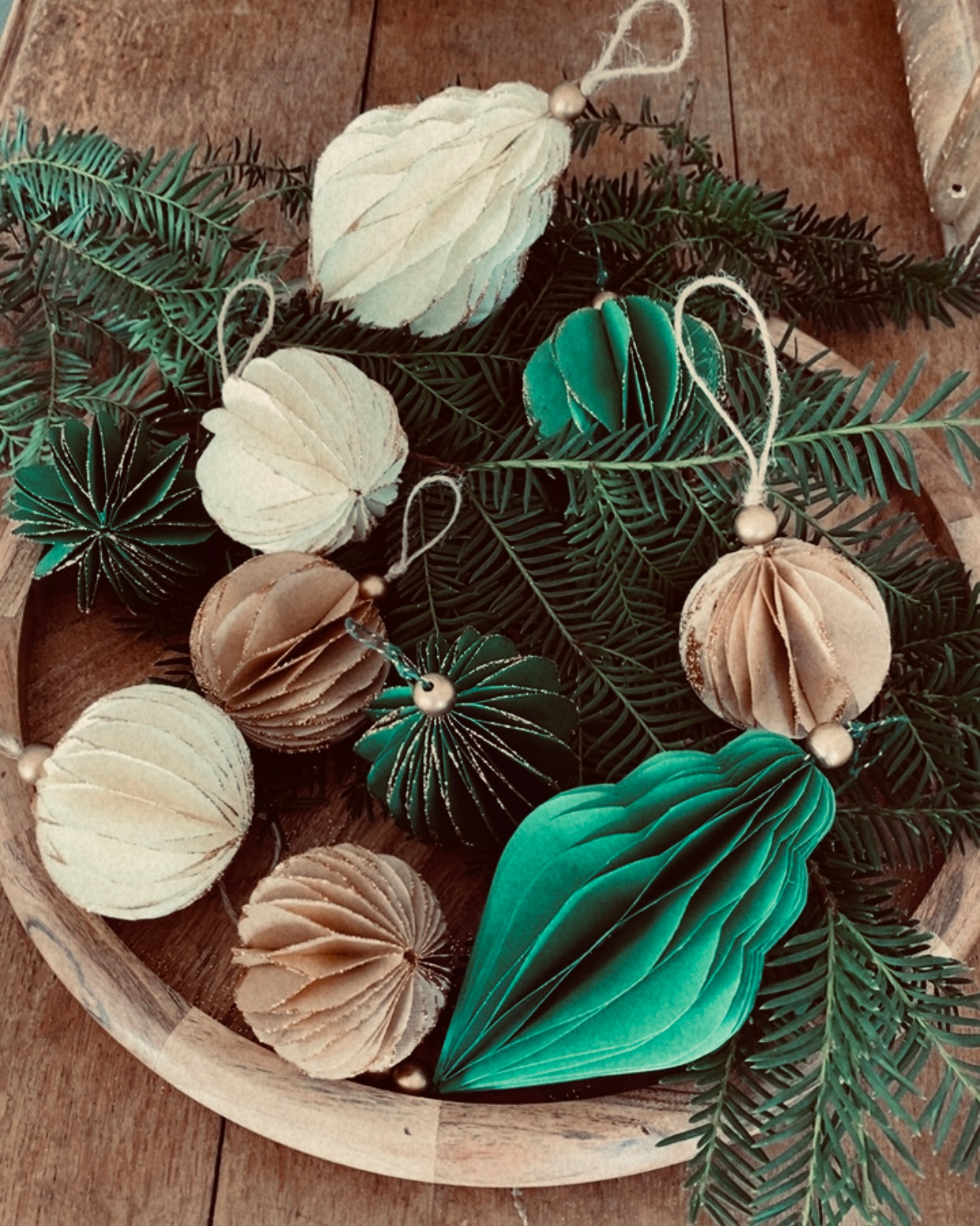 Décoration de Noël artisanale en papier, suspendue par une cordelette naturelle avec une perle dorée. La boule présente une forme élégamment travaillée en plusieurs couches découpées à la main, avec de légères touches de paillettes dorées qui soulignent ses contours. L’ornement, de teinte écru douce, est tenu dans une main, mettant en valeur son relief, sa finesse et son allure poétique, idéale pour décorer un sapin ou une guirlande de fête. ninona